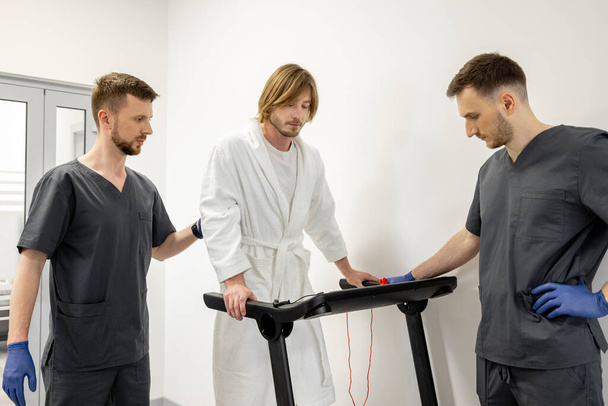 Two rehabilitation specialists help a patient walk on a treadmill during rehabilitation. Trauma treatment and medical support concept - Photo, image