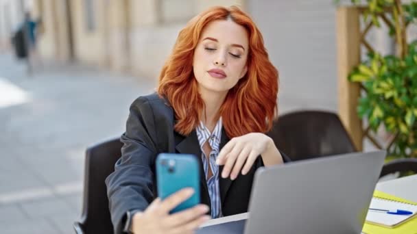 Νεαρή κοκκινομάλλα επιχειρηματίας χρησιμοποιώντας φορητό υπολογιστή κάνει selfie από το smartphone στη βεράντα καφετέρια - Πλάνα, βίντεο