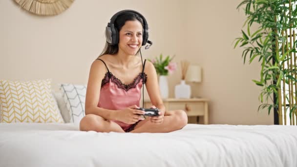 Joven hermosa mujer hispana jugando video juego sentado en la cama sonriendo en el dormitorio - Metraje, vídeo