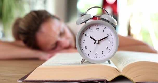 Alarm clock on book and woman sleeping in background. Education time concept of learning and loss of motivation - Metraje, vídeo
