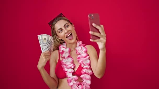 Junge blonde Frau im Bikini hält Dollars bei Videoanruf vor isoliertem roten Hintergrund - Filmmaterial, Video