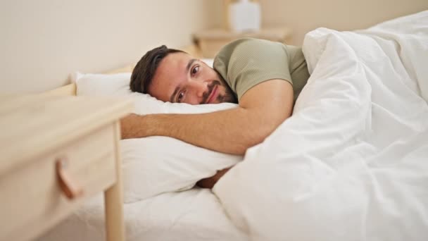 Νεαρός Ισπανός άντρας χαμογελάει με αυτοπεποίθηση ξαπλωμένος στο κρεβάτι στην κρεβατοκάμαρα - Πλάνα, βίντεο