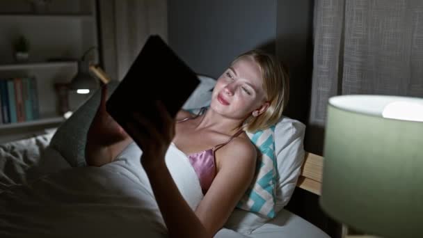 Giovane donna bionda utilizzando touchpad sdraiato sul letto in camera da letto - Filmati, video