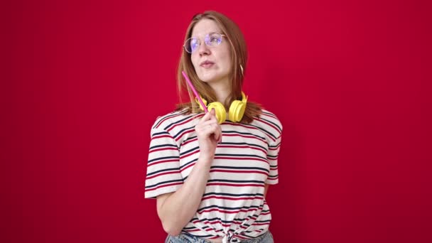 Junge blonde Frau mit Kopfhörern, die mit zweifelndem Gesichtsausdruck auf den isolierten roten Hintergrund zeigt - Filmmaterial, Video