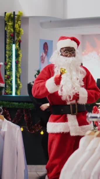 Vídeo vertical Dolly en la toma de alegre gerente vestido como Santa Claus en Navidad adornan tienda de ropa centro comercial. Supervisor jingling campanas de Navidad y saludar a los clientes en la moda temática de vacaciones - Imágenes, Vídeo