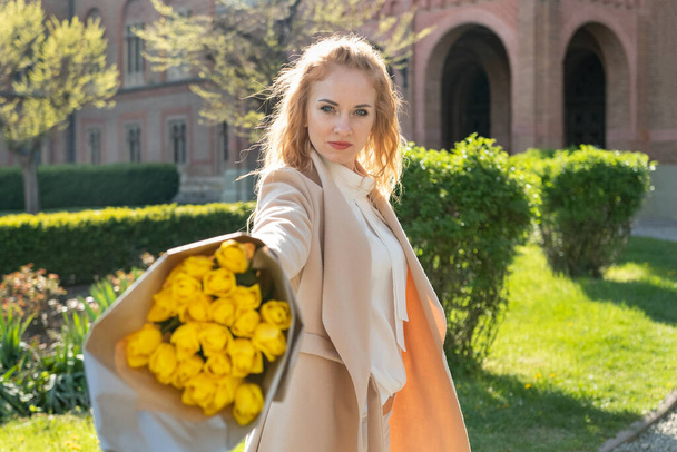 Eine blonde junge Frau mit einem Strauß gelber Blumen spaziert an einem sonnigen Frühlingstag durch die Stadt - Foto, Bild