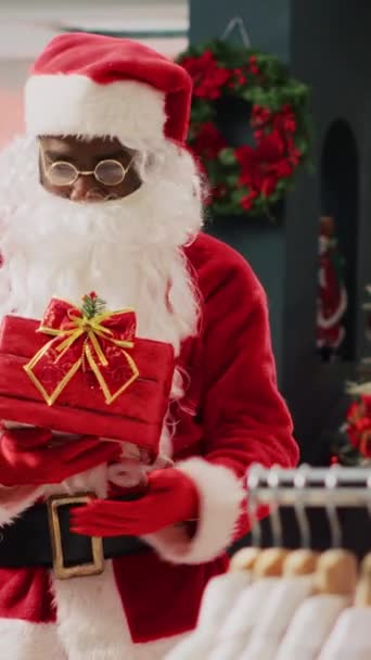 Vertikální video Zaměstnanec na sobě Santa Claus oblečení v obchodě zdobí vánoční oblečení, zve zákazníky k účasti na vánoční tomboly soutěže s cílem vyhrát propagační cenu během zimy - Záběry, video