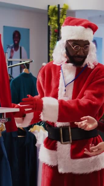 Vertical video Manager portant le costume du Père Noël autour du centre commercial boutique de mode pour répandre la joie des vacances. Superviseur en costume à thème festif montrant gai client pièce de vêtement rouge - Séquence, vidéo