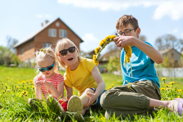 I bambini felici si divertono in cortile di casa, la giornata estiva calda. Fratelli e sorelle seduti sull'erba in denti di leone depositati - Foto, immagini