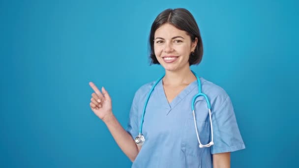 Junge schöne hispanische Ärztin lächelt und zeigt zur Seite über isolierten blauen Hintergrund - Filmmaterial, Video