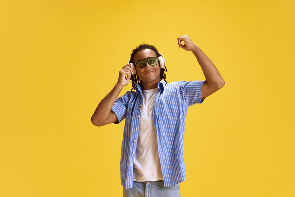 Joven hombre alegre en gafas de sol y camisa a cuadros escuchando música en auriculares sobre fondo de estudio amarillo. Concepto de juventud, emociones humanas, estilo de vida, moda, expresiones faciales, anuncio - Foto, Imagen