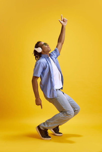 Full-length image of young man in sunglasses and casual clothes listening to music in headphones and dancing against yellow studio background. Concept of youth, human emotions, lifestyle, fashion, ad - Foto, immagini