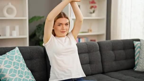 Jonge blonde vrouw ontspannen met handen op het hoofd zitten op de bank thuis - Video