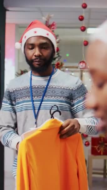 同僚が情報を得るのに役立つ間,顧客のために優雅なブレーザーを持って来る従業員の垂直ビデオ追跡ショット. クリスマスオルネイトでクライアントに衣服を提示する小売アシスタント - 映像、動画
