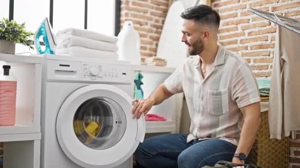 Jovem hispânico sentado no chão começando máquina de lavar roupa olhando assistir na lavanderia - Filmagem, Vídeo