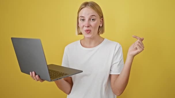 jonge blonde vrouw wijzen naar laptop verrast over geïsoleerde gele achtergrond - Video