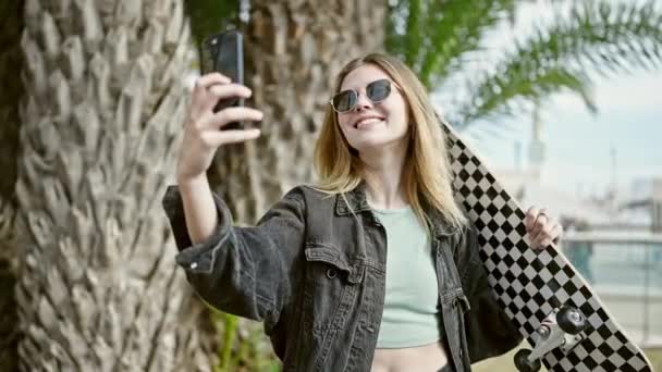 Молодая блондинка с коньками делает селфи на смартфоне, улыбаясь в парке. - Кадры, видео