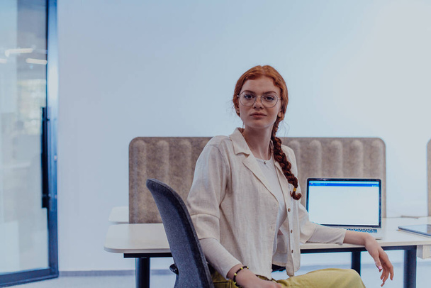 A modern businesswoman with striking orange hair sitting confidently in a contemporary office, accompanied by her laptop, embodying professionalism, creativity, and success in a technology-driven - Photo, image