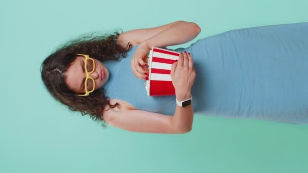 Nadšená mladá žena ve 3D brýle jíst popcorn a sledovat zajímavé televizní seriál, sportovní hra, film, on-line sociální média filmový obsah. Krásná dospělá dívka izolované na modrém pozadí studia. Svisle - Záběry, video