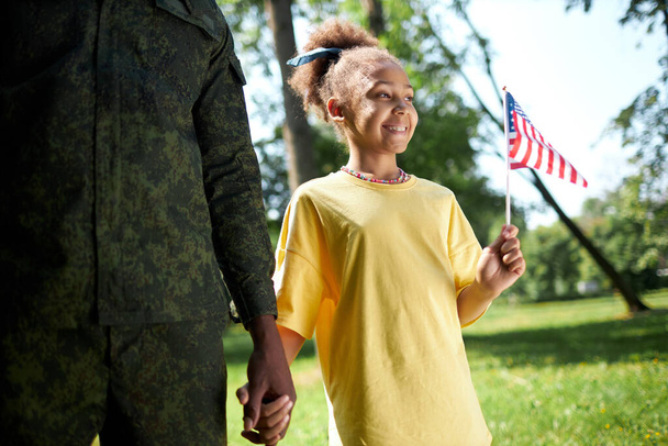Petite fille afro-américaine avec drapeau américain marchant avec son père en uniforme militaire à l'extérieur - Photo, image
