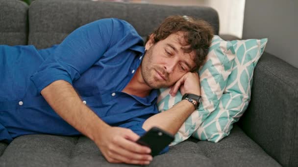 Fiatal spanyol férfi használ okostelefon fekvő kanapén fáradt otthon - Felvétel, videó