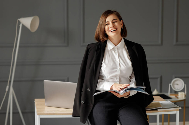 Lachende zakenvrouw draagt een wit shirt, een zwart jasje op haar schouders en een zwarte jeans houdt een tijdschrift in haar handen, leunend op een bureaublad met een laptop op tafel en een witte lamp achter haar op de achtergrond van een versierde grijze wal - Foto, afbeelding