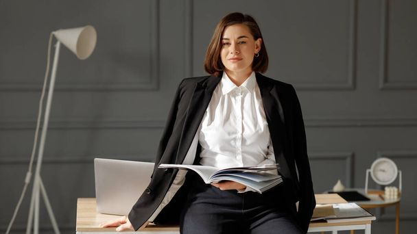 Серйозно бізнес-леді носить білу сорочку, чорна куртка на плечах і чорні джинси тримає в руках журнал, спираючись на стільницю з ноутбуком на столі і білою лампою за нею на тлі прикрашеного сірого кольору - Фото, зображення
