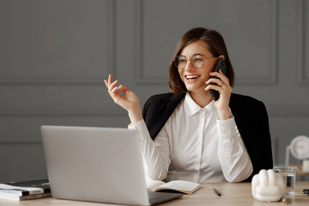 Een mooie zakenvrouw met een bril op haar ogen, met een wit shirt en een zwart jasje op haar schouders, zit achter een bureau voor een laptop en praat aan de telefoon in het kantoor - Foto, afbeelding