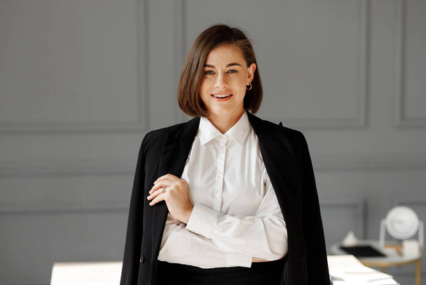 Glimlachende zakenvrouw met mooie make-up is het dragen van witte shirt en zwarte jas op schouders staat met armen gekruist in de voorkant van grijze kamer achtergrond - Foto, afbeelding