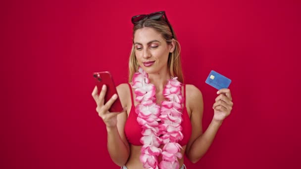 Молодая блондинка в бикини со смартфоном и кредиткой на изолированном красном фоне - Кадры, видео