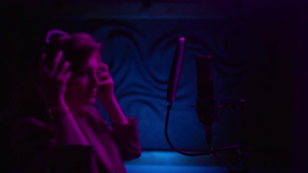 Egy tehetséges énekes lány portréja, aki új dalt énekel az audió stúdióban. Nyugodt nő professzionális fejhallgatóval és mikrofonnal új albumot készít pop dalokkal a stúdióban neon fényben - Felvétel, videó