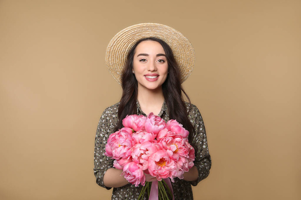 Beautiful young woman in straw hat with bouquet of pink peonies against light brown background - Foto, imagen