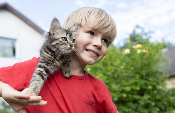 смугастий пухнастий кошеня сидить на плечі милого хлопчика 7 років, дивлячись в одному напрямку. день кота. щасливе дитинство. Ніжна позитивна атмосфера спілкування між тваринами і людьми - Фото, зображення