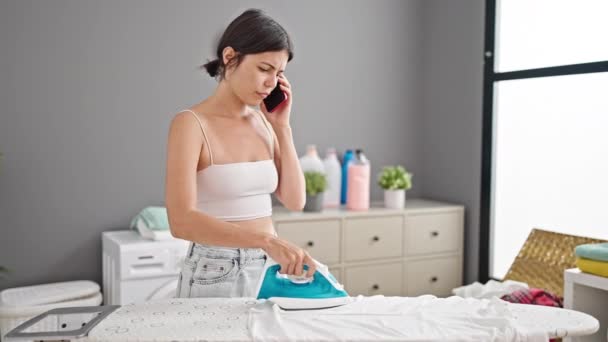 Young beautiful hispanic woman talking on smartphone ironing clothes at laundry room - Footage, Video