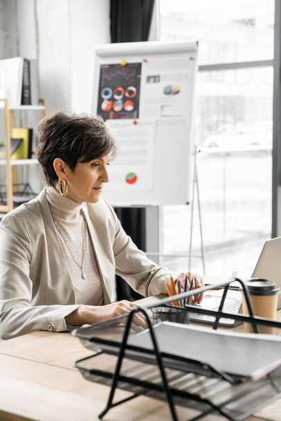 middle aged businesswoman working on laptop near coffee to go in office, confidence and experience - Photo, image