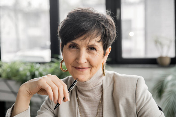 mature woman entrepreneur with pen looking at camera in office, experience, professional headshot - Foto, imagen