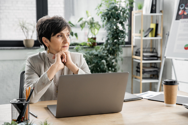 thoughtful businesswoman sitting near laptop and looking away in modern office, executive, ambitious - Photo, image