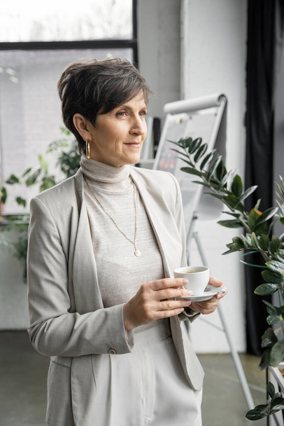 confident and experienced middle aged entrepreneur with coffee cup looking away in modern office - Photo, image