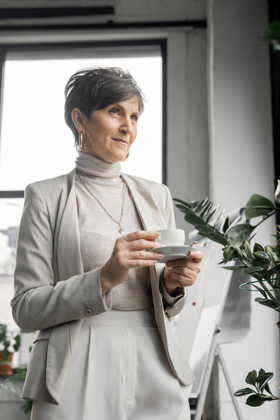 positive and dreamy middle aged businesswoman with coffee cup looking away in modern office - Foto, afbeelding