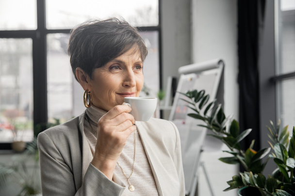 επιτυχημένη μεσήλικη γυναίκα, εταιρικός διευθυντής, πίνοντας καφέ και κοιτάζοντας μακριά στο γραφείο - Φωτογραφία, εικόνα