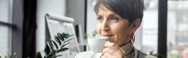 pleased mature woman, executive manager, drinking coffee and looking away in modern office, banner - Photo, Image