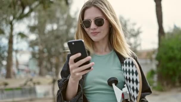 Giovane donna bionda che tiene il pattino utilizzando lo smartphone al parco - Filmati, video