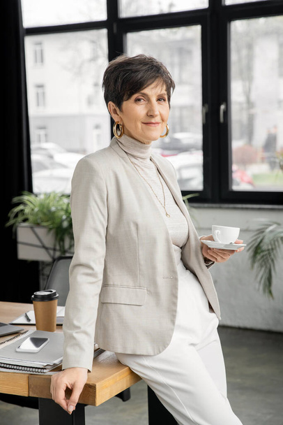 exitosa mujer de negocios de mediana edad con taza de café sonriendo a la cámara en la oficina moderna, disparo en la cabeza - Foto, imagen