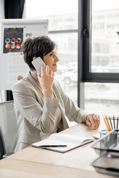 optimista gerente de mediana edad hablando en el teléfono móvil cerca de portátil en el lugar de trabajo en la oficina moderna - Foto, Imagen