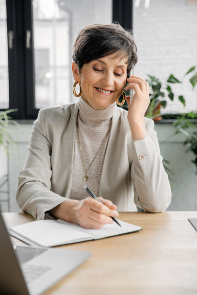 Gülümseyen orta yaşlı iş kadını akıllı telefonla konuşuyor ve modern ofiste deftere yazıyor. - Fotoğraf, Görsel