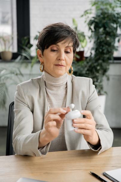 středního věku podnikatelka drží pouzdro na sluchátka se sluchátky na pracovišti v moderní kanceláři - Fotografie, Obrázek