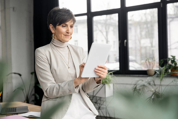 femme d'affaires d'âge moyen positive avec tablette numérique travaillant dans un bureau contemporain - Photo, image