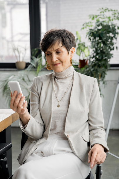 smiling middle aged businesswoman networking on smartphone at workplace in modern office - Photo, Image