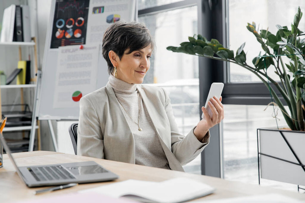 mujer de negocios de mediana edad con teléfono inteligente cerca del ordenador portátil y rotafolio borroso con gráficos en la oficina - Foto, Imagen