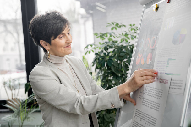 positive middle aged manager working with business analytics on flip chart in modern office - Photo, Image